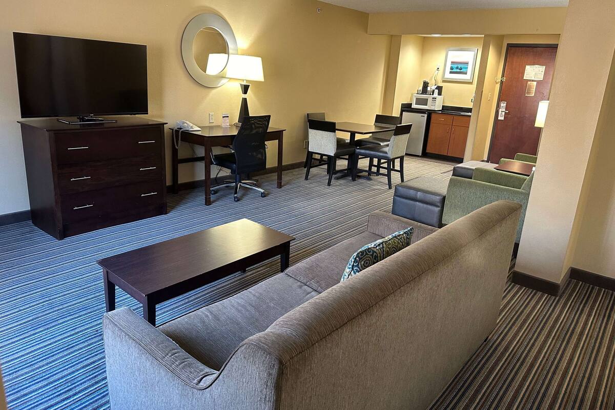 A hotel room with a sofa, a coffee table, a TV, a desk with a chair, and a kitchenette in the background.