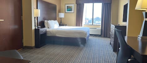 A hotel room with a bed, a desk, a chair, a lamp, a nightstand, a window with curtains, and a painting on the wall.