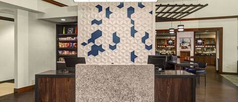 A reception area with a modern design, featuring a central counter with a marble top, a decorative wall with geometric patterns, and a wooden ceiling.