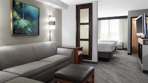 A hotel room with a sofa, a small wooden table, a bed, a TV, and a painting on the wall. - Hyatt Place Boise/Towne Square (Boise)