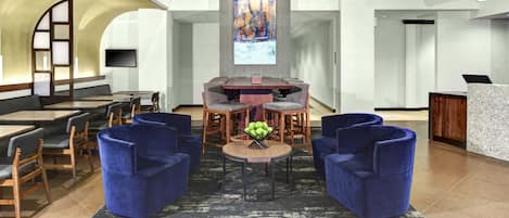 A lobby with a reception desk, seating area with blue chairs, a round table, and a large abstract painting on the wall.