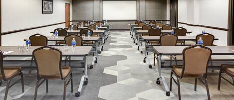 A conference room with rows of tables and chairs, equipped with water bottles and presentation screens.