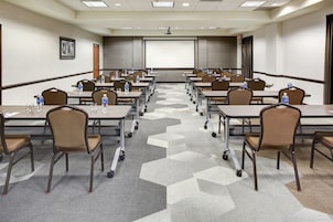 Un salón de conferencias con filas de mesas y sillas, equipado con botellas de agua y pantallas para presentaciones.