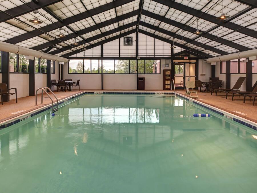 An indoor swimming pool with clear water, surrounded by lounge chairs and tables, under a high ceiling with skylights.