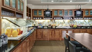A modern kitchen with wooden cabinets, a glass display case, and a buffet area with various food items. - Hyatt Place Boise/Towne Square (Boise)
