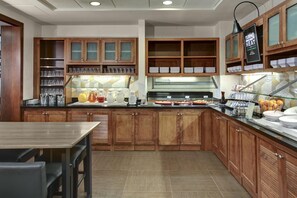 A modern kitchen with wooden cabinets, a glass display case, and a buffet counter with various food items. - Hyatt Place Boise/Towne Square (Boise)