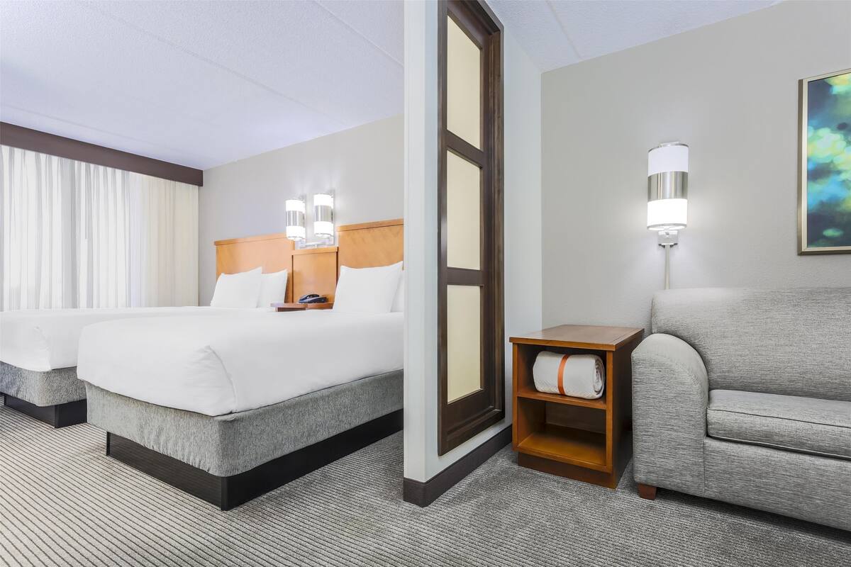 A hotel room with two beds, a sofa, a wooden nightstand, and a wall-mounted lamp.