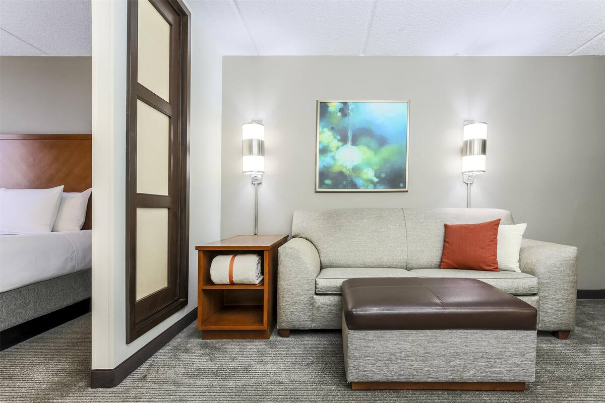 A hotel room with a bed, a sofa, an ottoman, a side table, and two wall-mounted lamps.