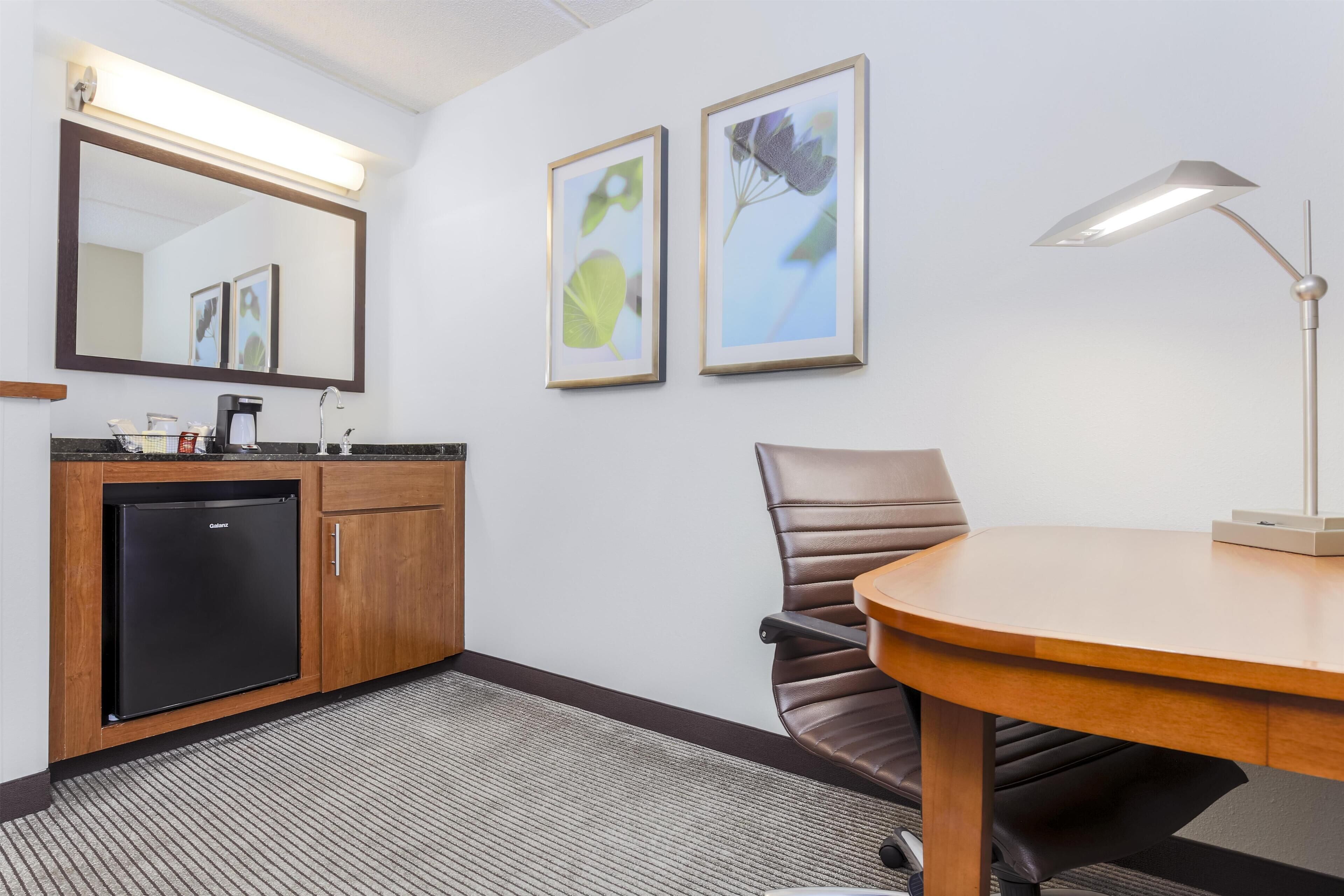 A hotel room with a wooden desk, a chair, a black refrigerator, a sink, and two framed pictures on the wall.