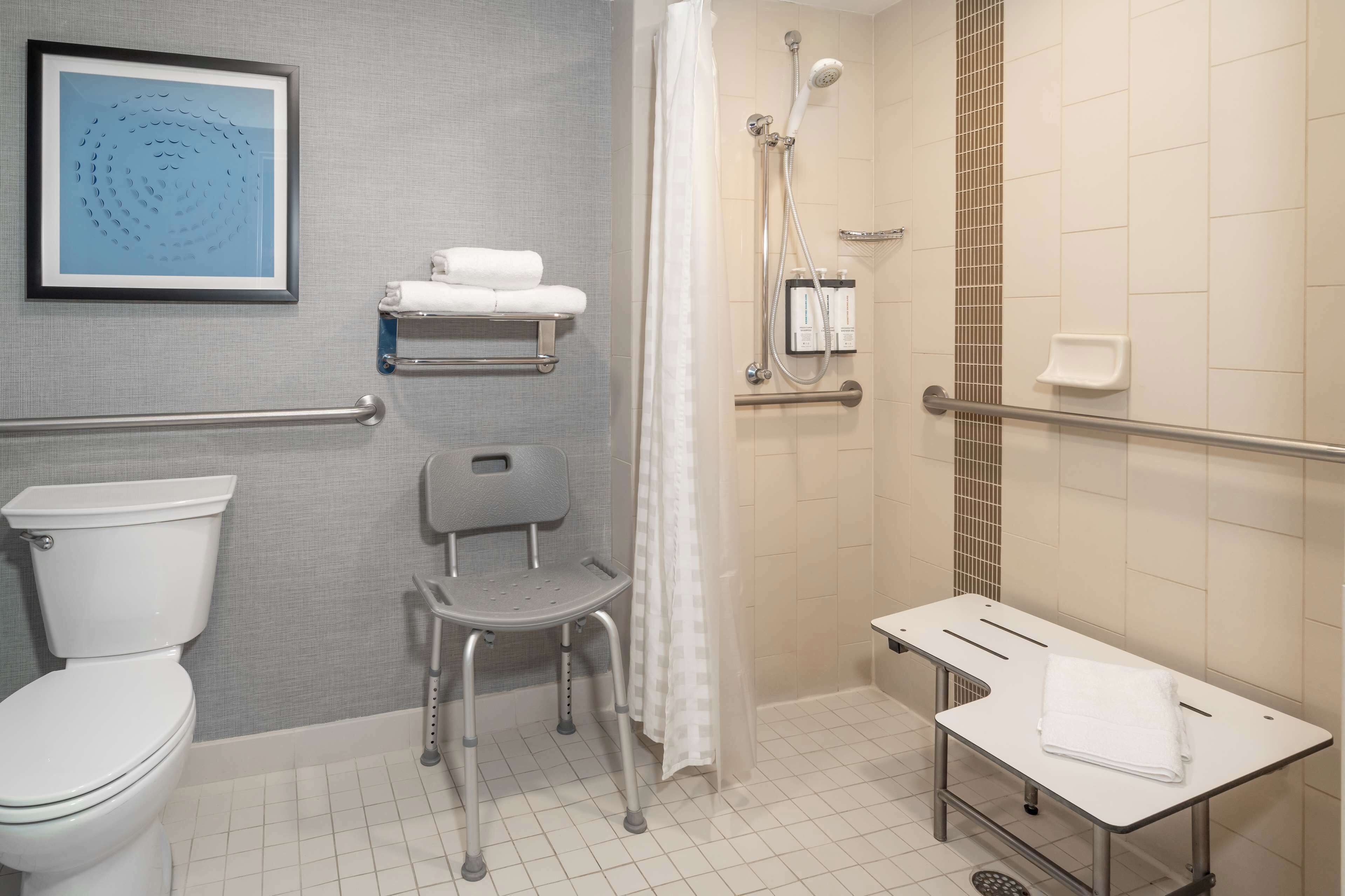 A wheelchair-accessible bathroom with a toilet, a shower with a handheld showerhead, a grab bar, a towel rack, and a bench.
