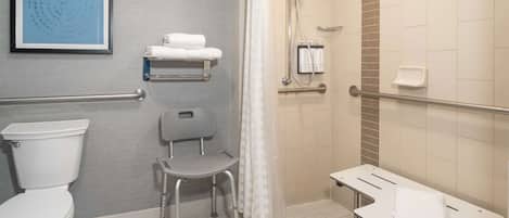 A wheelchair-accessible bathroom with a toilet, a shower with a handheld showerhead, a grab bar, a towel rack, and a bench.