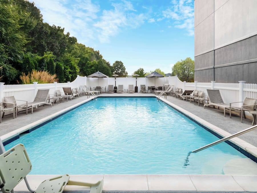 A rectangular pool with lounge chairs and umbrellas, surrounded by a white fence and greenery.