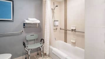 A bathroom with a bathtub, a toilet, a handrail, a shower curtain, a grab bar, a wheelchair, and a towel rack.