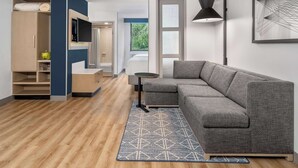 A modern living room with a grey sectional sofa, a small round table, a blue patterned rug, a TV mounted on a wall, and a large window with a view of greenery. - Hyatt Place Mystic (Mystic)