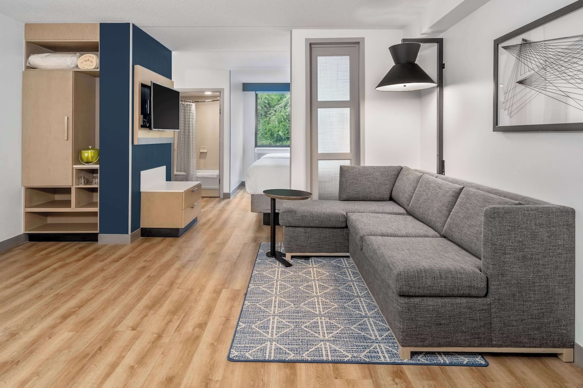 Un salon moderne avec un canapé d’angle gris, une petite table ronde, un tapis bleu à motifs, un téléviseur fixé au mur et une grande fenêtre donnant sur un paysage verdoyant.
