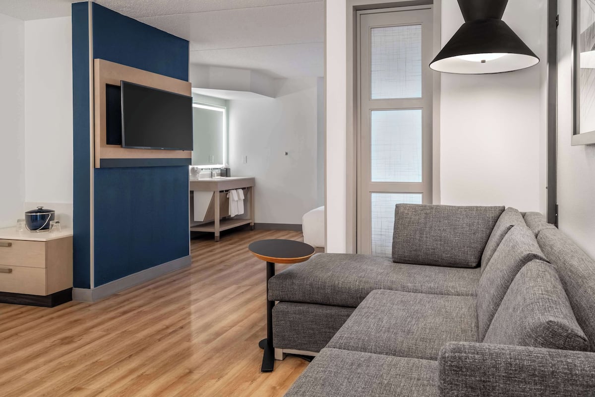Un salon moderne avec un canapé gris, une petite table ronde, un téléviseur fixé sur un mur bleu qui sert de point d’attraction, et une grande fenêtre avec des stores.
