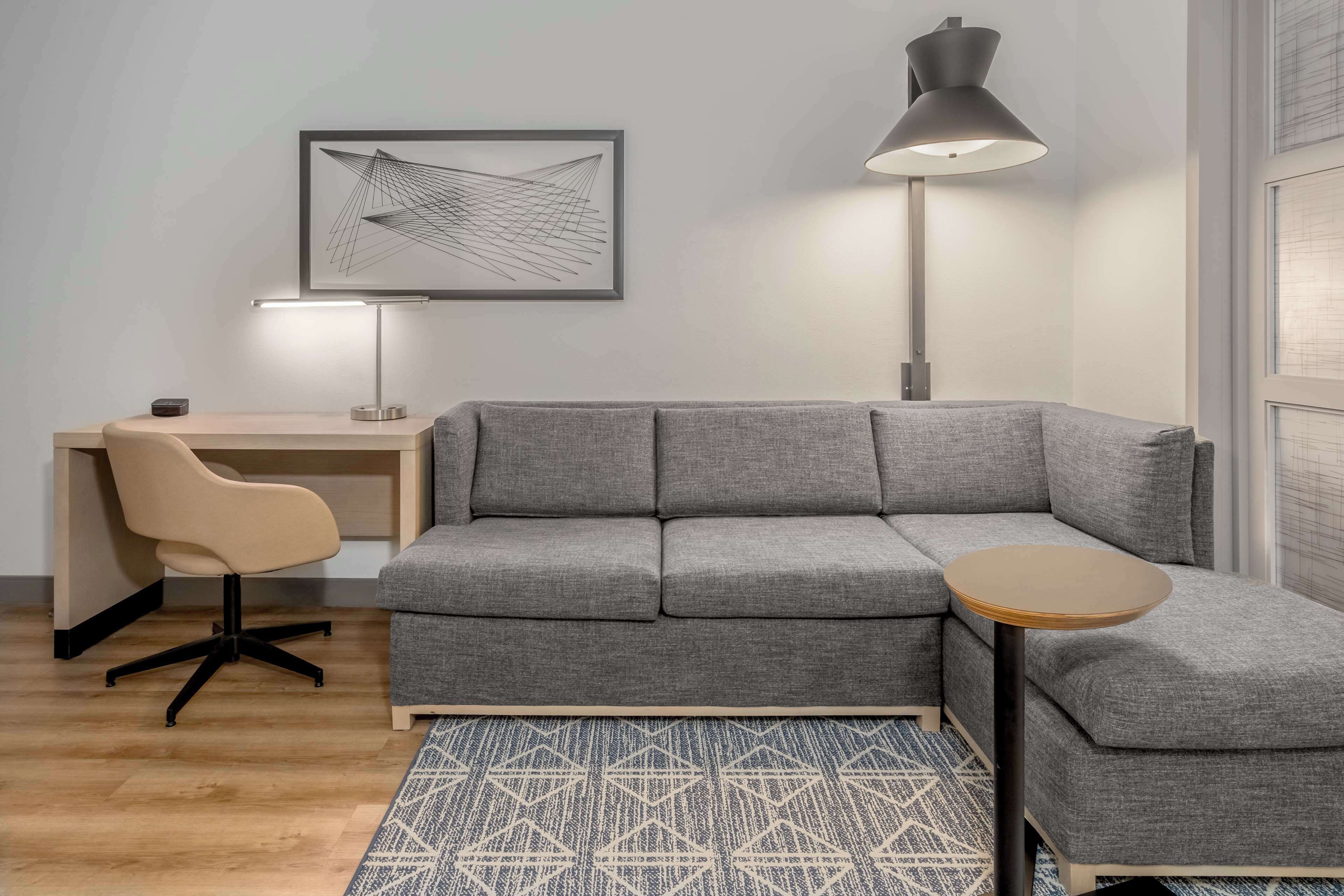 A modern living room with a grey sectional sofa, a small round table, a desk with a chair, a floor lamp, and a geometric patterned rug.
