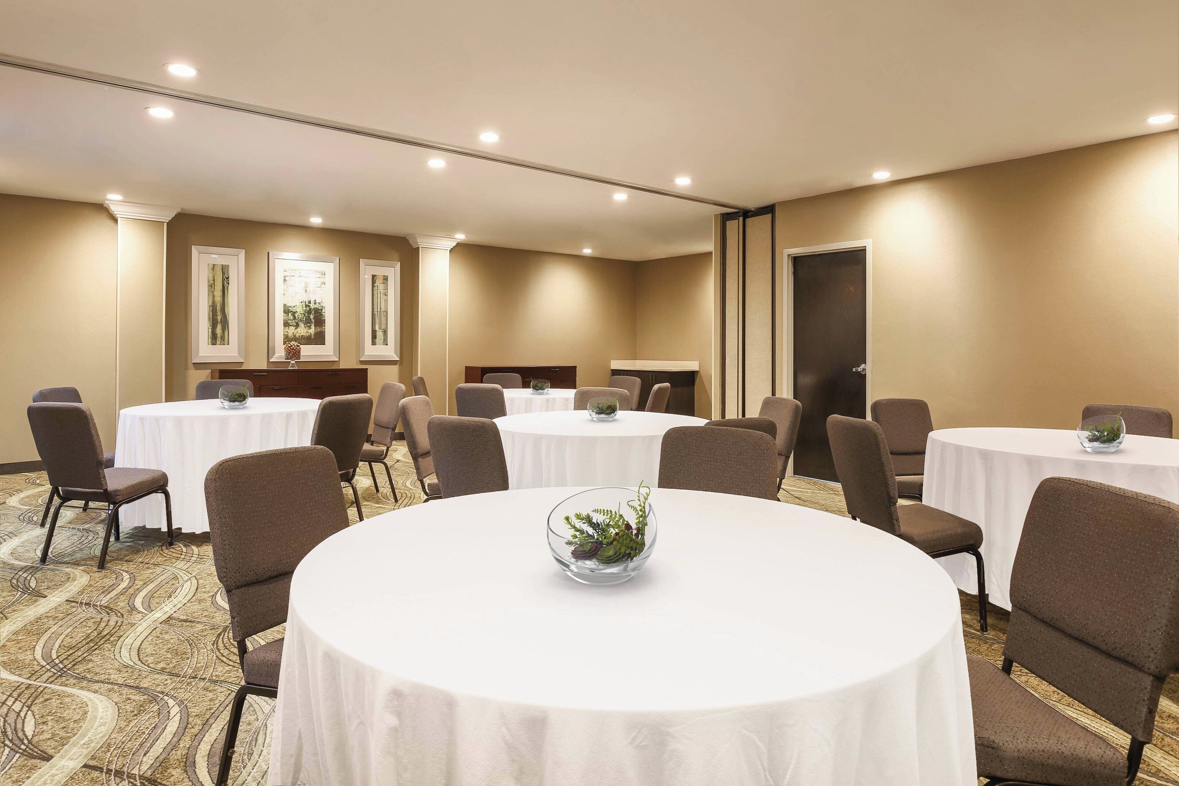 A conference room with multiple round tables, each with a white tablecloth and a glass bowl with greenery. The room has beige walls and a carpeted floor.