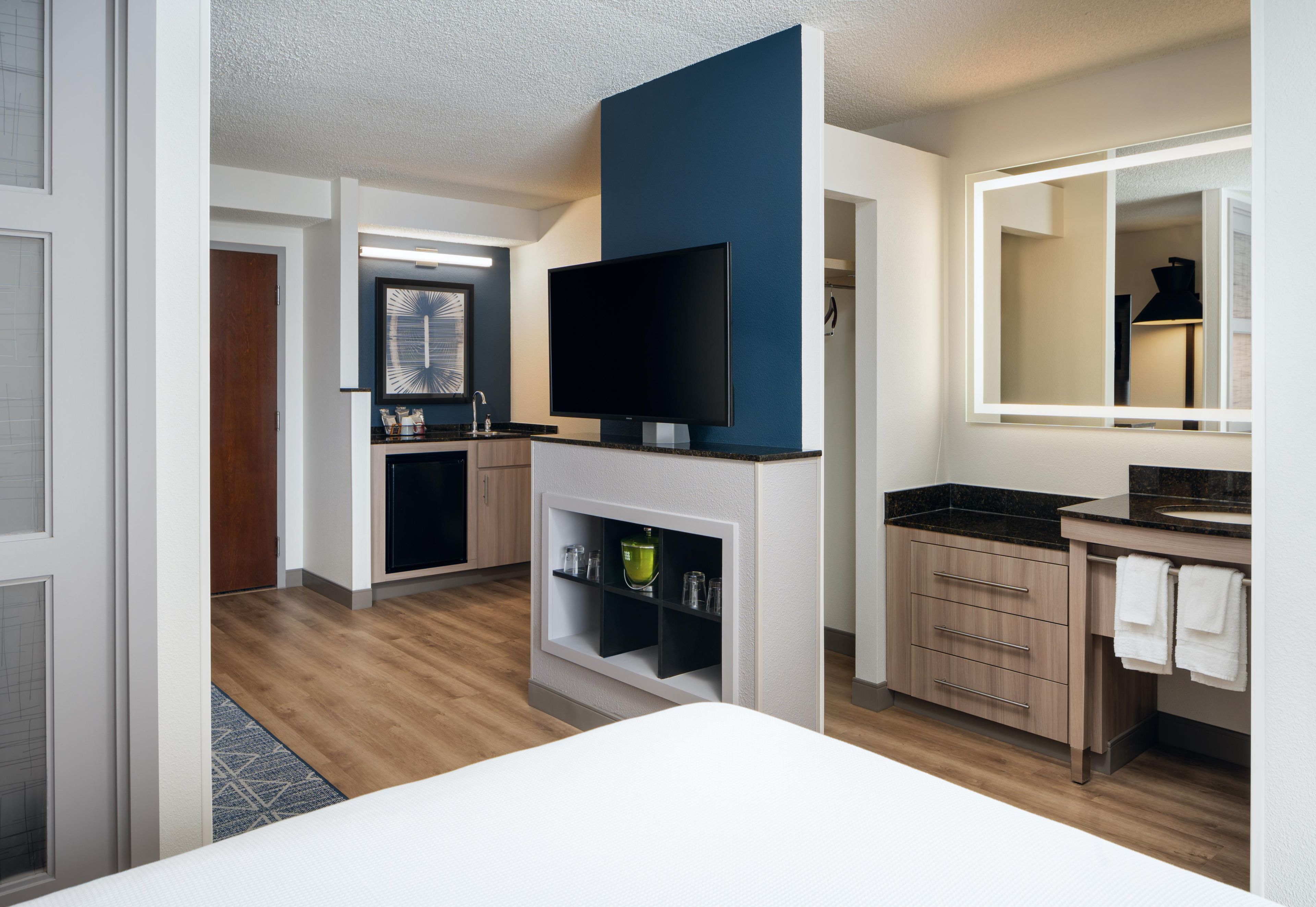 A modern hotel room with a flat-screen TV mounted on a wall, a wooden cabinet with a sink, and a mirror above the counter.