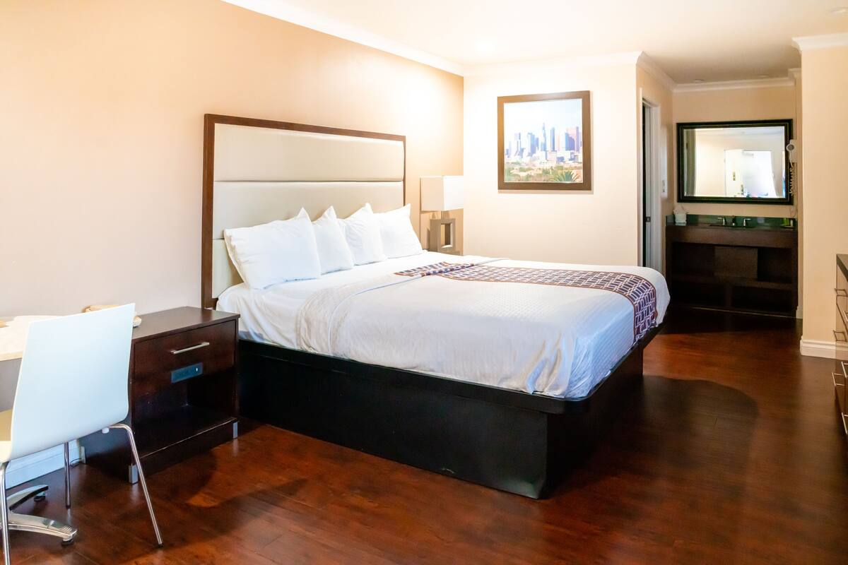 A hotel room with a large bed, a desk, a chair, a nightstand, a mirror, and a painting on the wall.