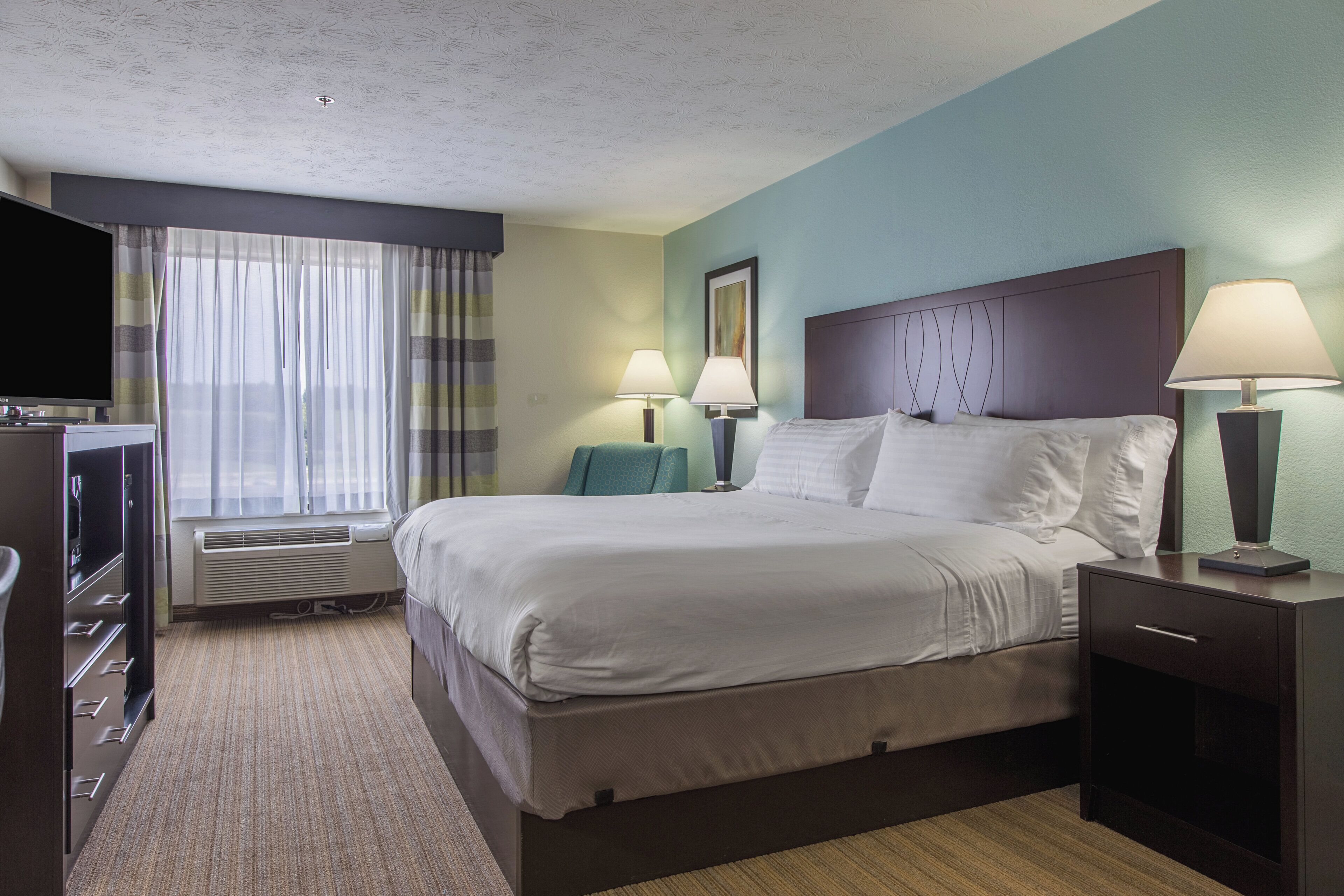 Une chambre d’hôtel avec un lit, deux lampes de chevet, une télévision, un bureau et une fenêtre avec des rideaux.
