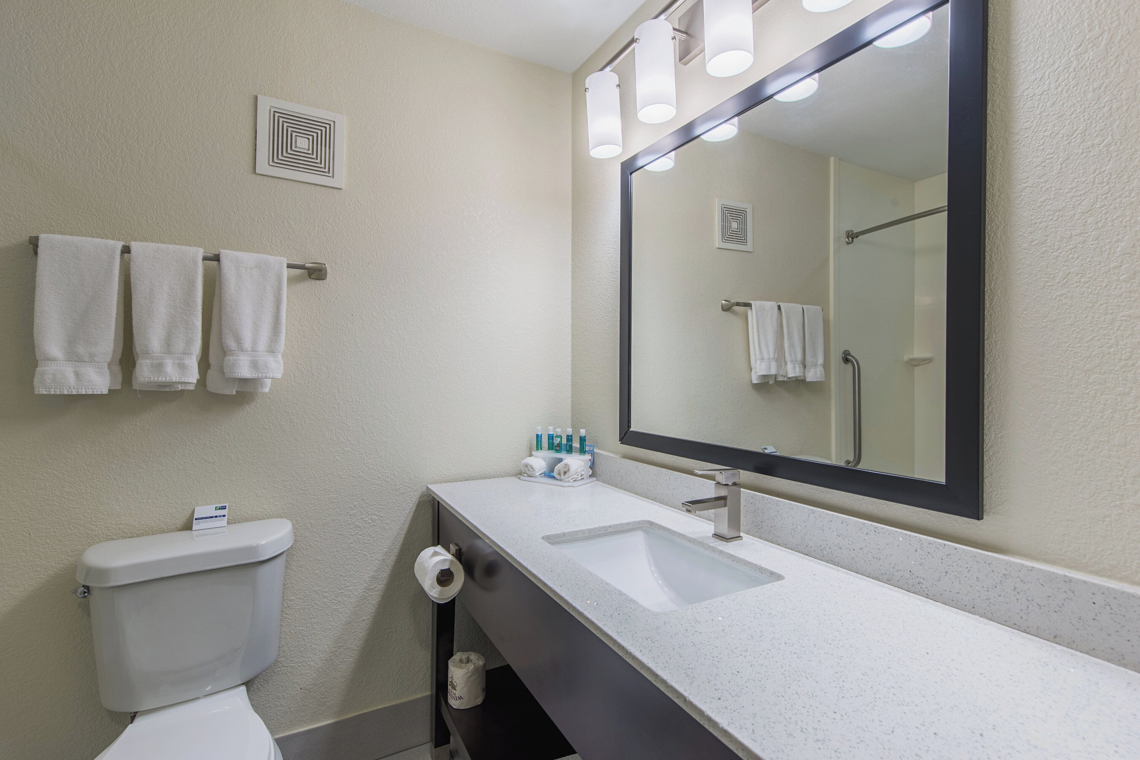 Een badkamer met een wit wastafel, een spiegel en een toilet. Handdoeken hangen aan een rek en er is een rol toiletpapier aan de muur.