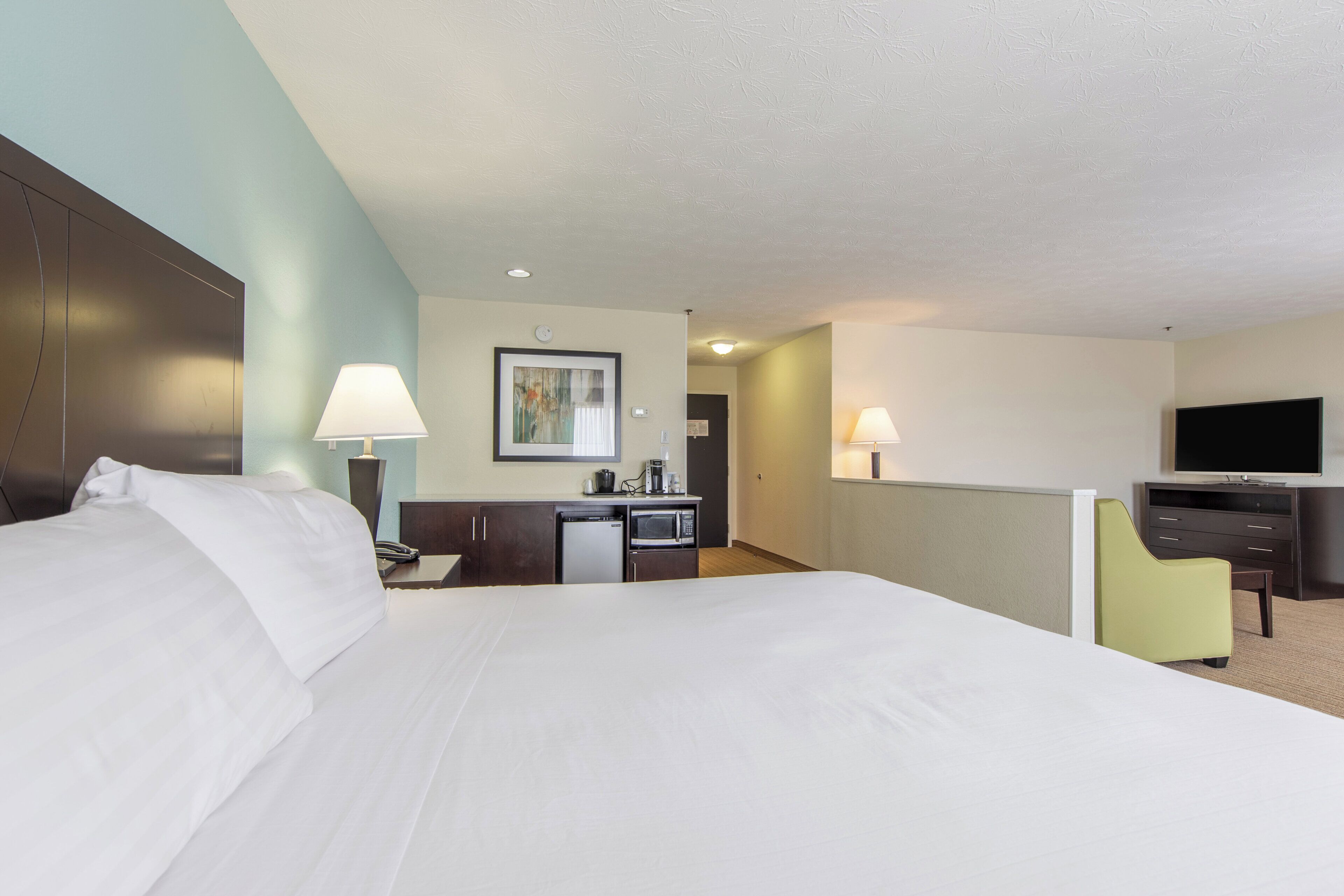 A hotel room with a large bed, a desk with a lamp, a television, and a small kitchenette.