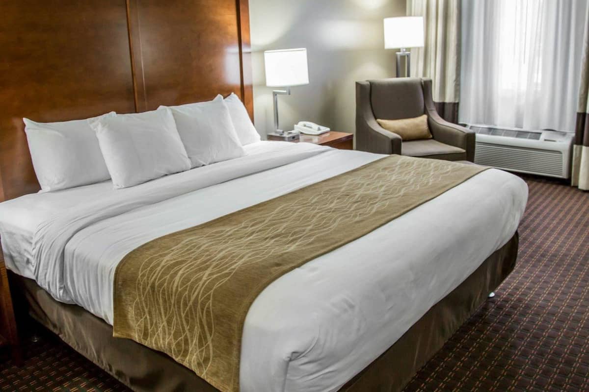 A hotel room with a large bed, a chair, a desk with a lamp, and an air conditioning unit.