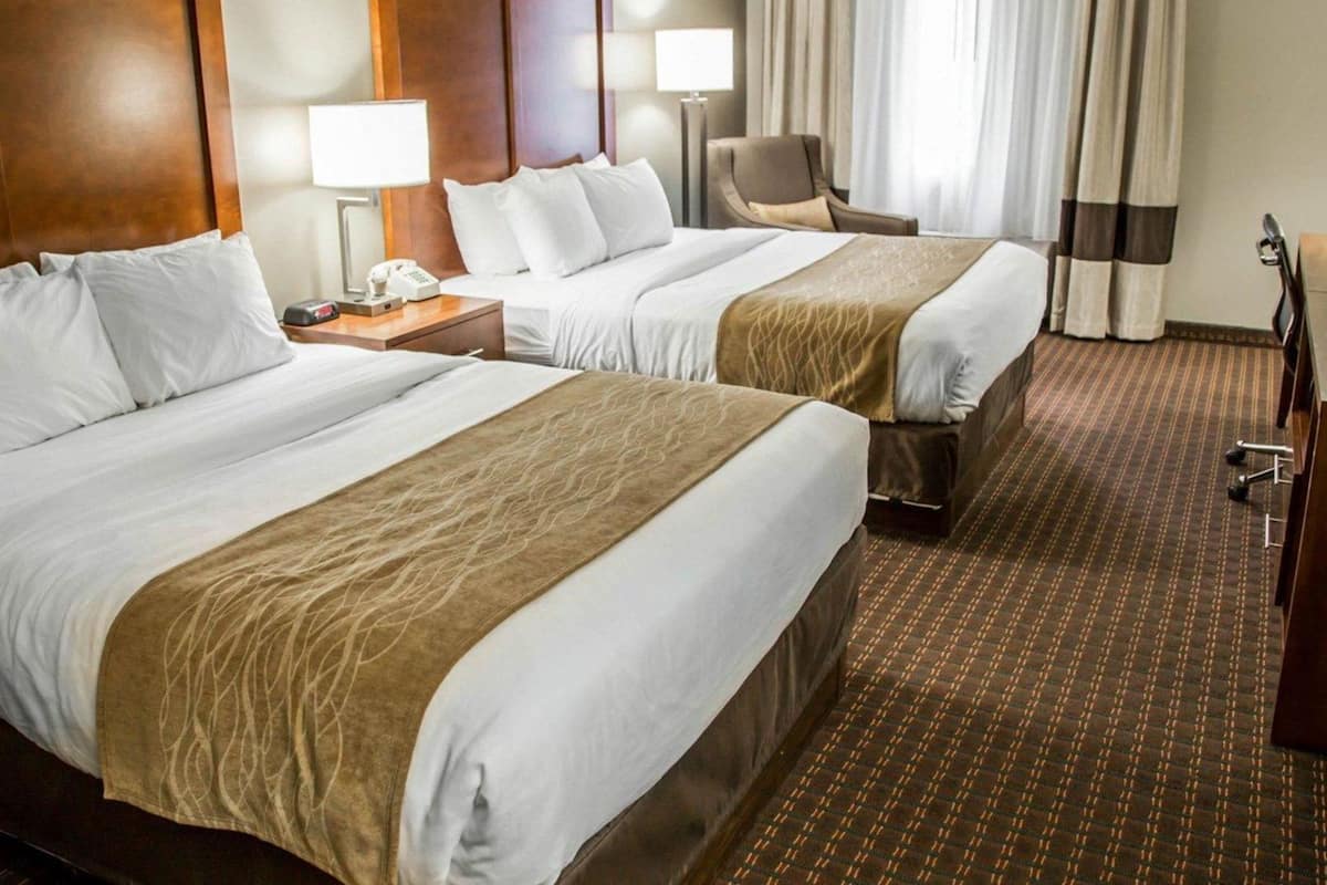 A hotel room with two beds, a desk, a chair, a lamp, and a large window with curtains.
