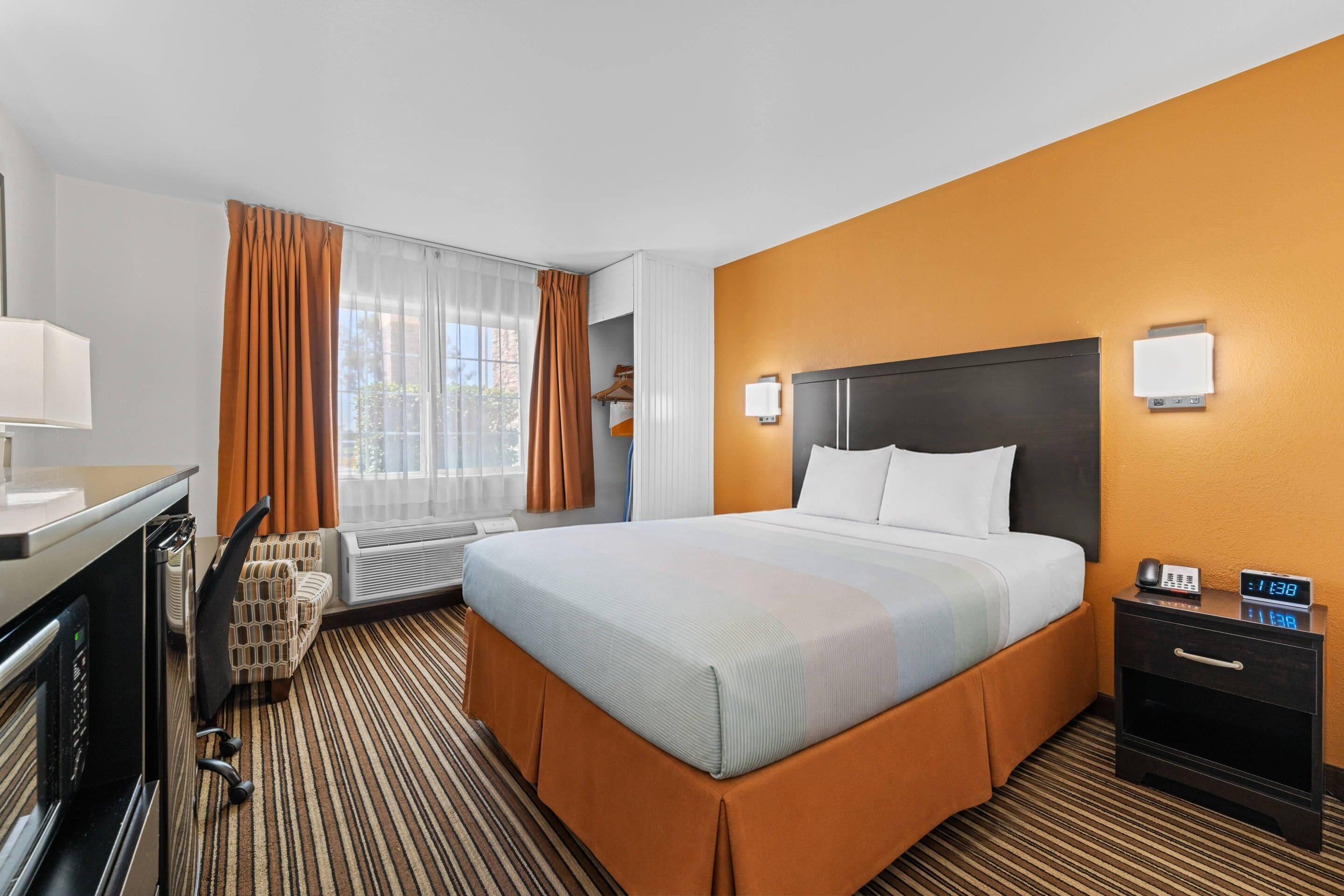 Une chambre d’hôtel comprenant un lit, un bureau, une chaise, une table de chevet avec une horloge et une fenêtre avec des rideaux.