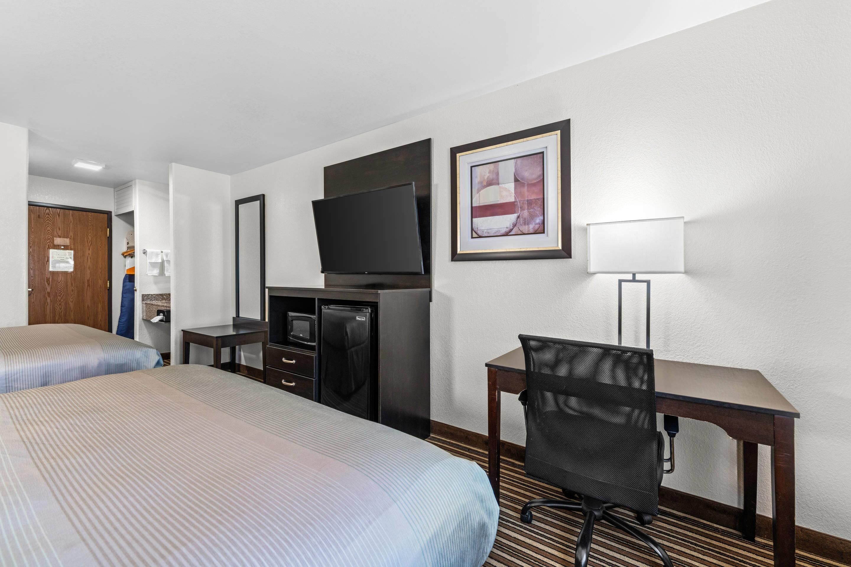 A hotel room with two beds, a desk, a television, a framed picture, and a lamp.