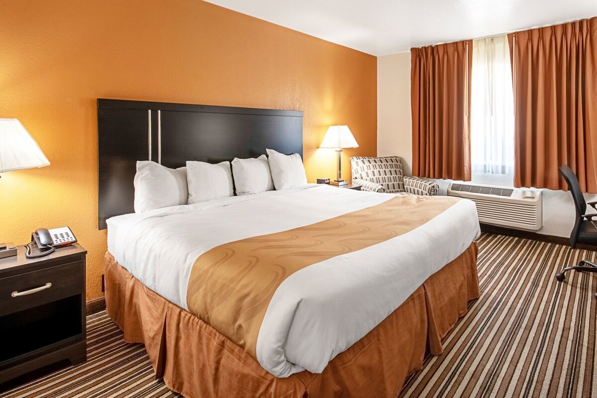 A hotel room with a large bed, two bedside lamps, a desk with a telephone, an air conditioning unit, and a patterned carpet.