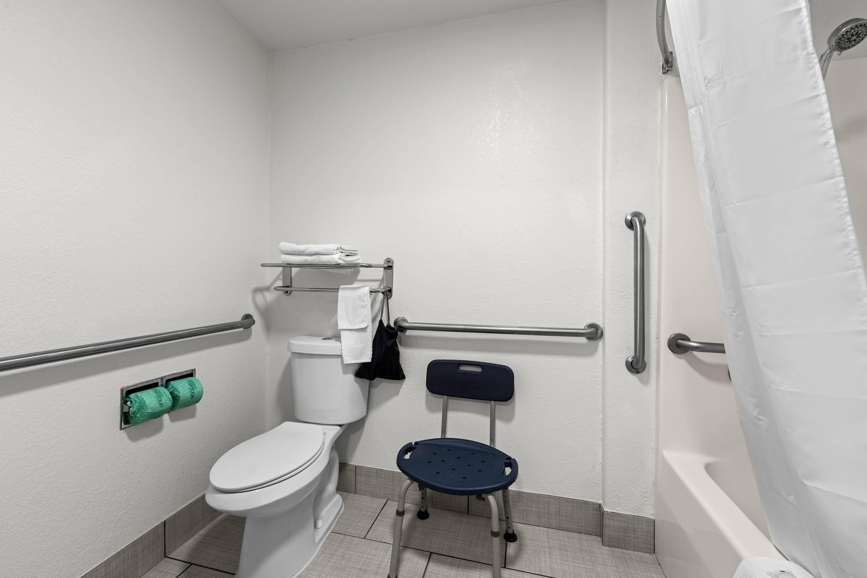 A bathroom with a toilet, a shower, a grab bar, a wheelchair-accessible chair, and a towel rack.