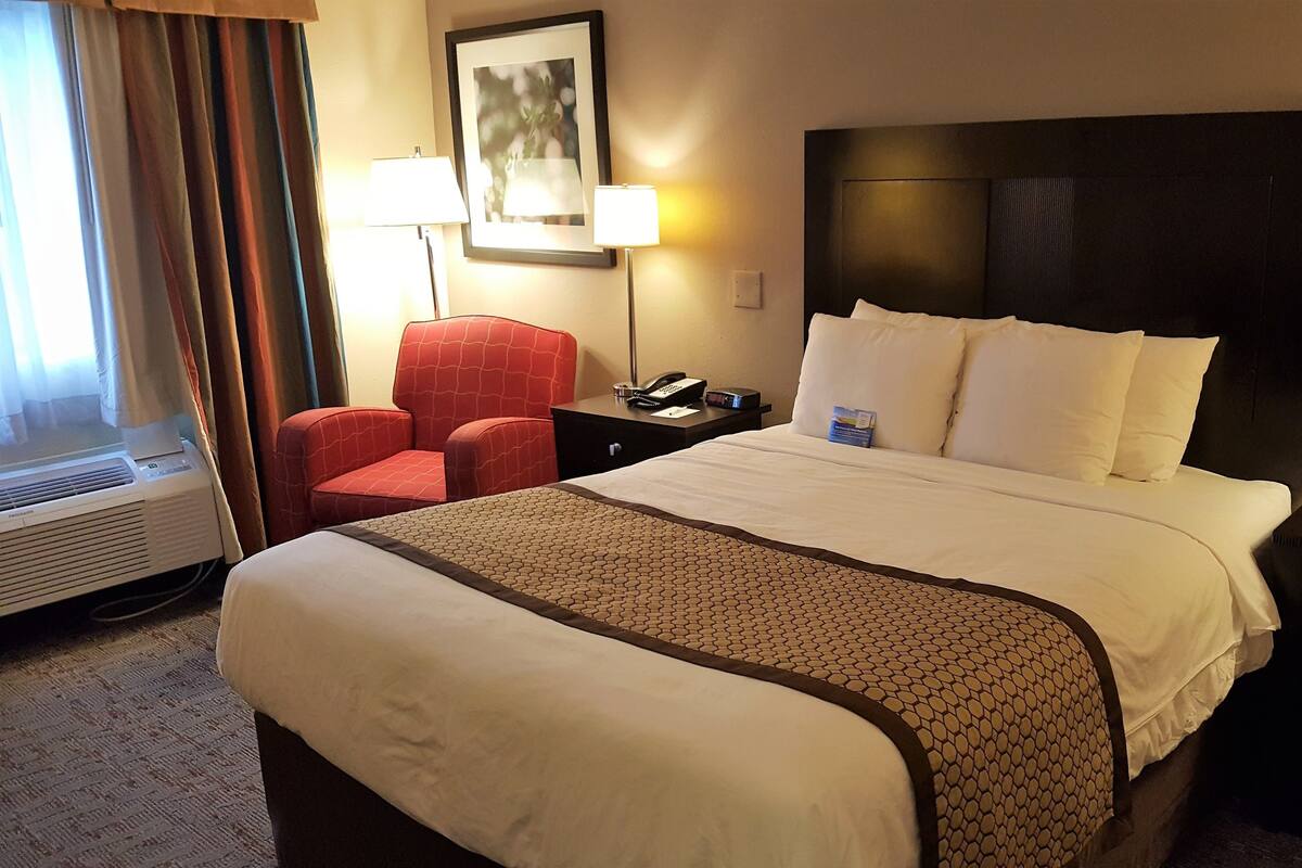 Une chambre d’hôtel avec un grand lit, un fauteuil rouge, un bureau avec un téléphone et une lampe, et un tableau accroché au mur.