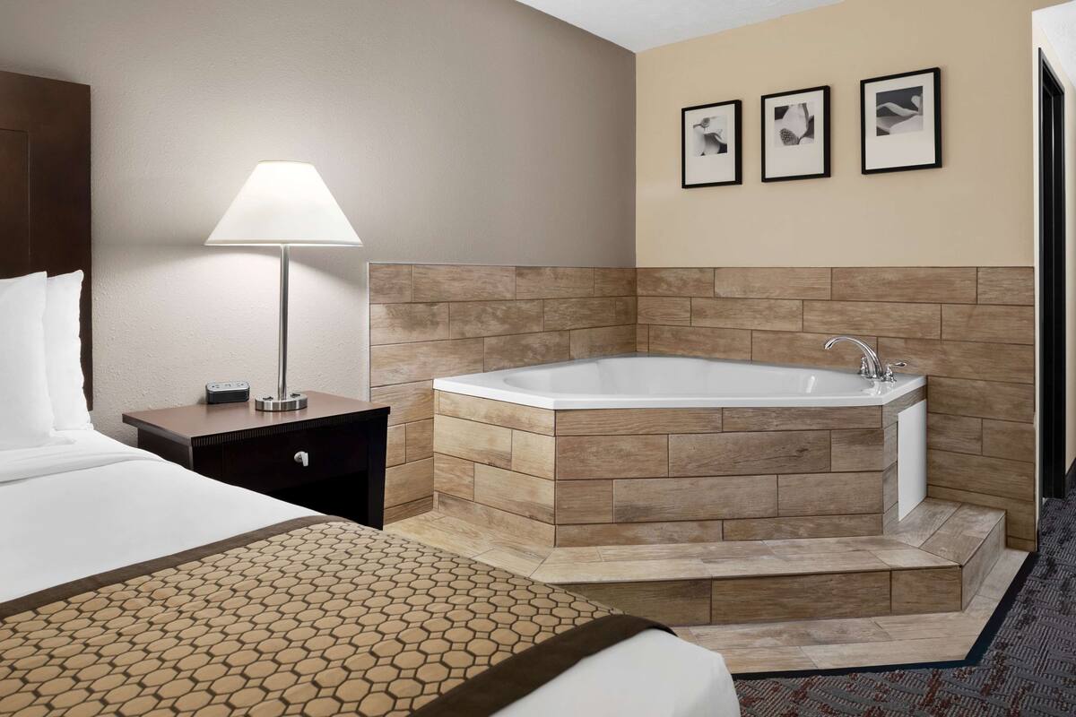 Une chambre d’hôtel avec un grand lit, une table de chevet avec une lampe, une baignoire d’angle et trois tableaux encadrés au mur.