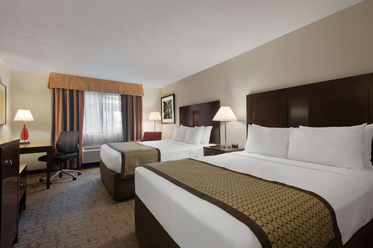 Une chambre d’hôtel avec deux lits, un bureau, une chaise, une lampe et une fenêtre avec des rideaux.