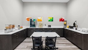 A modern kitchen with a central island, dark cabinetry, and a white countertop. There are two black chairs at the island, a coffee machine, and various food items displayed on the wall. - Quality Suites Houston NW Cy-Fair (Houston)