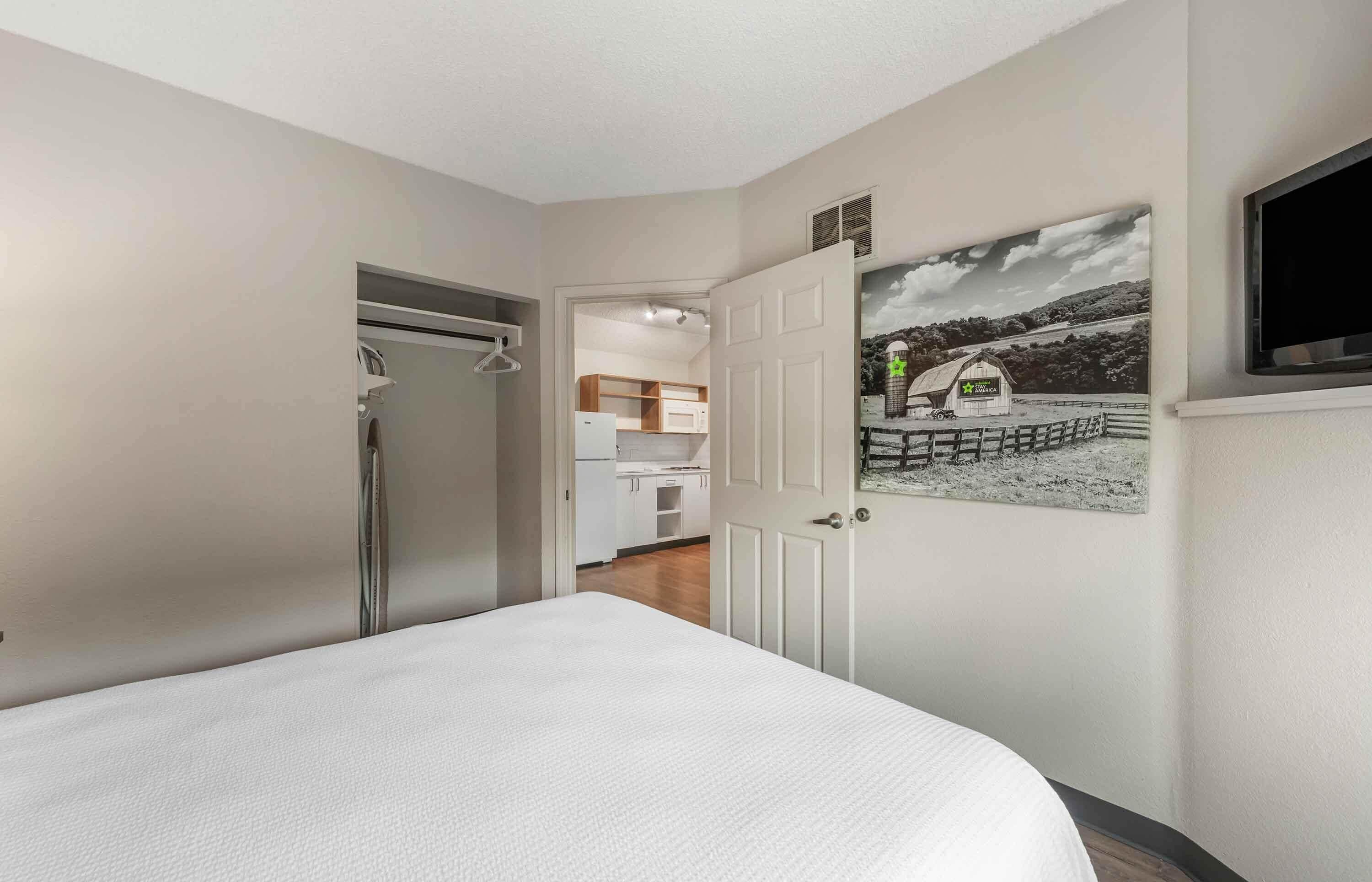 Un dormitorio con cama, una puerta que da a una cocina y un televisor montado en la pared.