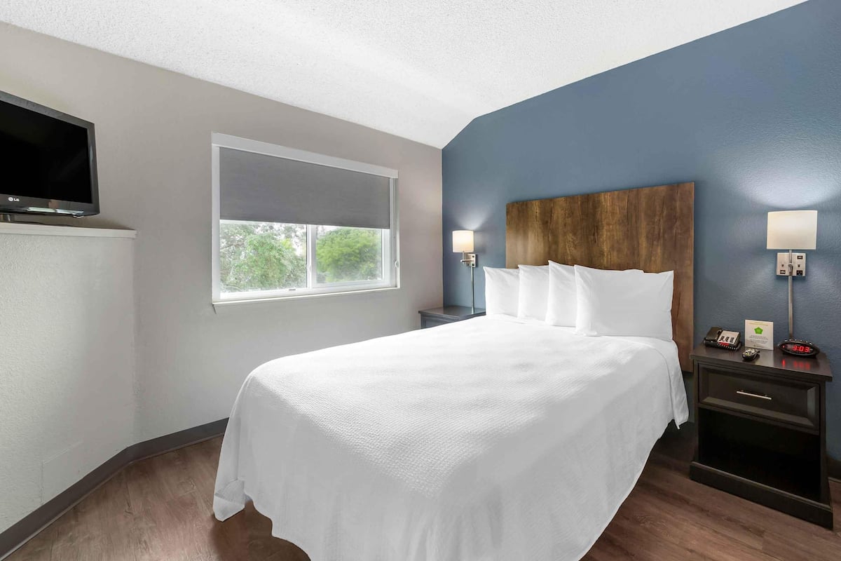 A hotel room with a large bed, a TV mounted on the wall, a window with blinds, a nightstand with a lamp, and a wall clock.