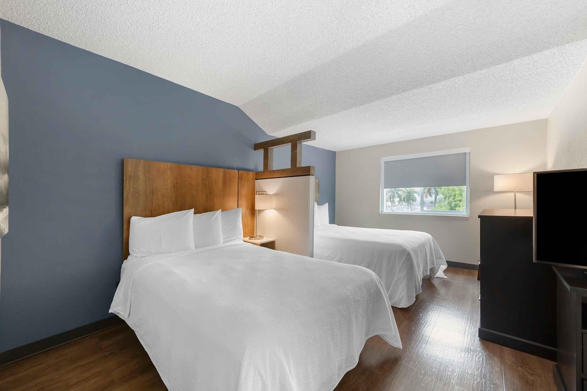 A hotel room with two beds, a wooden headboard, a window with a view, a desk, and a TV.