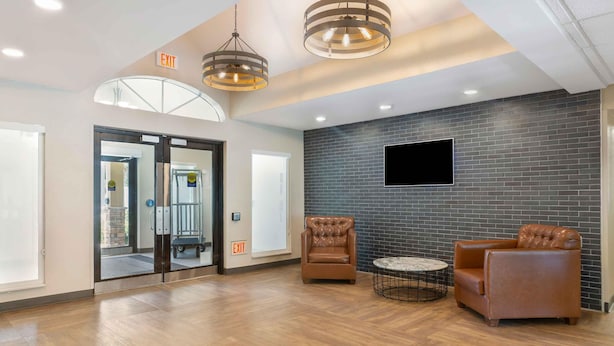 A lobby with a wooden floor, two leather armchairs, a round table, and a flat-screen TV mounted on a brick wall. - Extended Stay America Premier Suites - Miami - Airport - Miami Springs (Miami Springs)