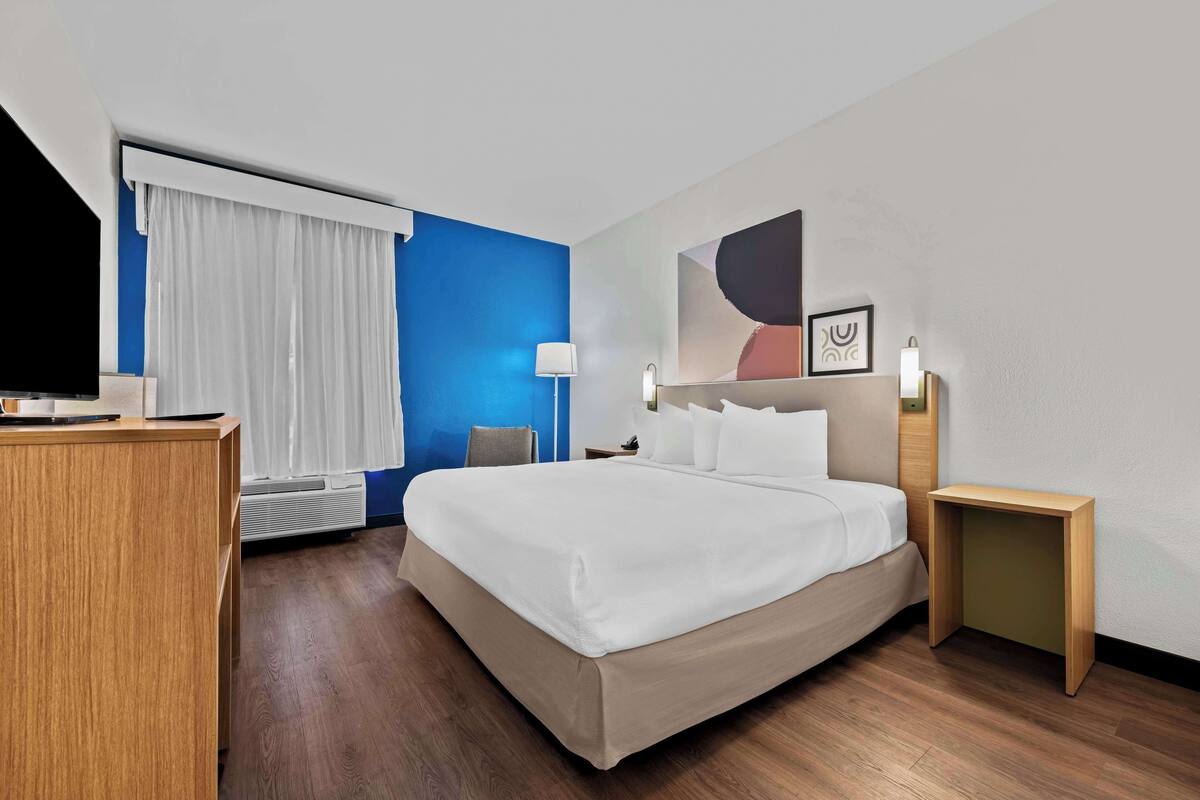 Habitación de hotel con cama, escritorio, silla, televisor, lámpara y una mesita.