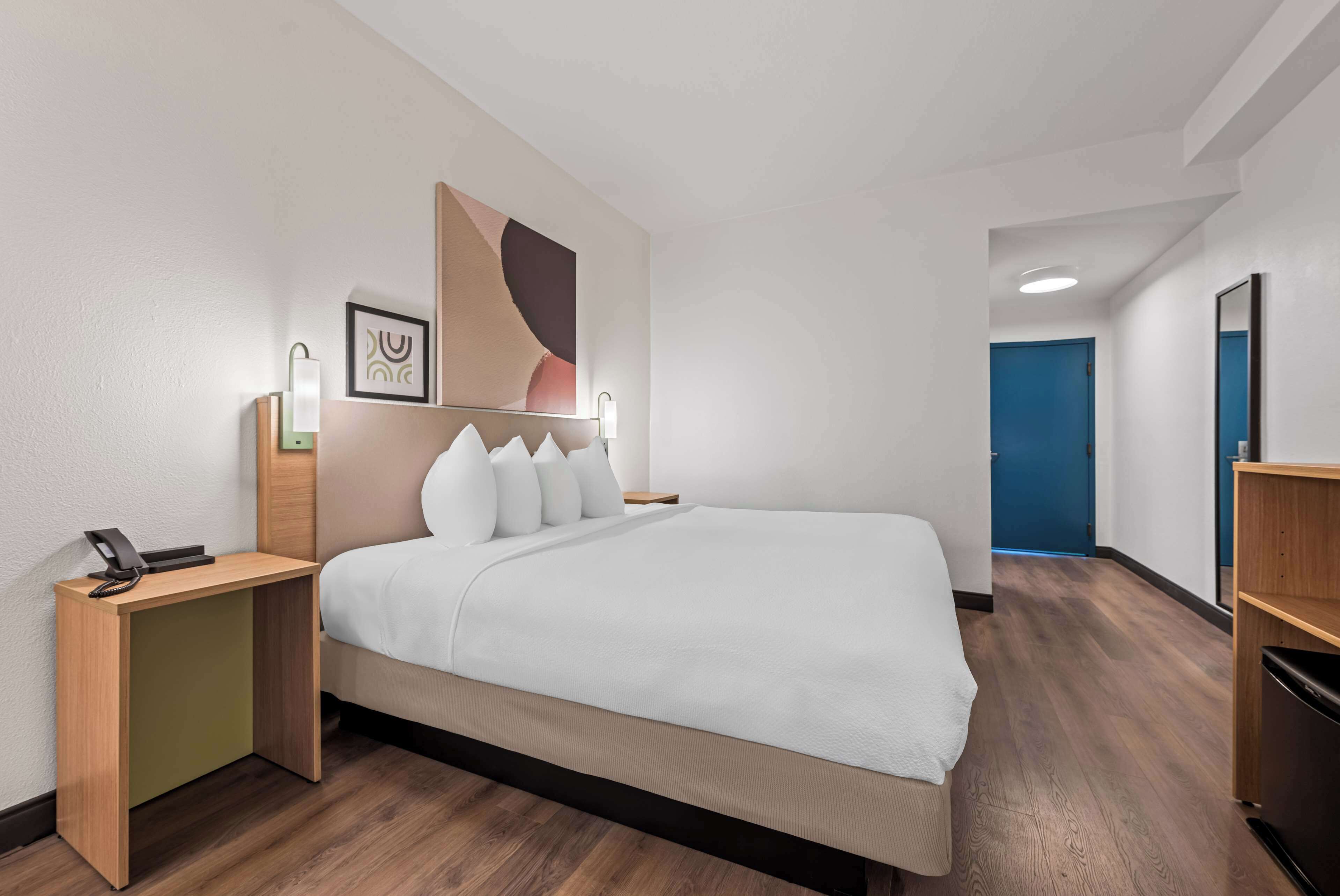 A hotel room with a large bed, a bedside table with a phone, a wall-mounted light, and a blue door in the background.
