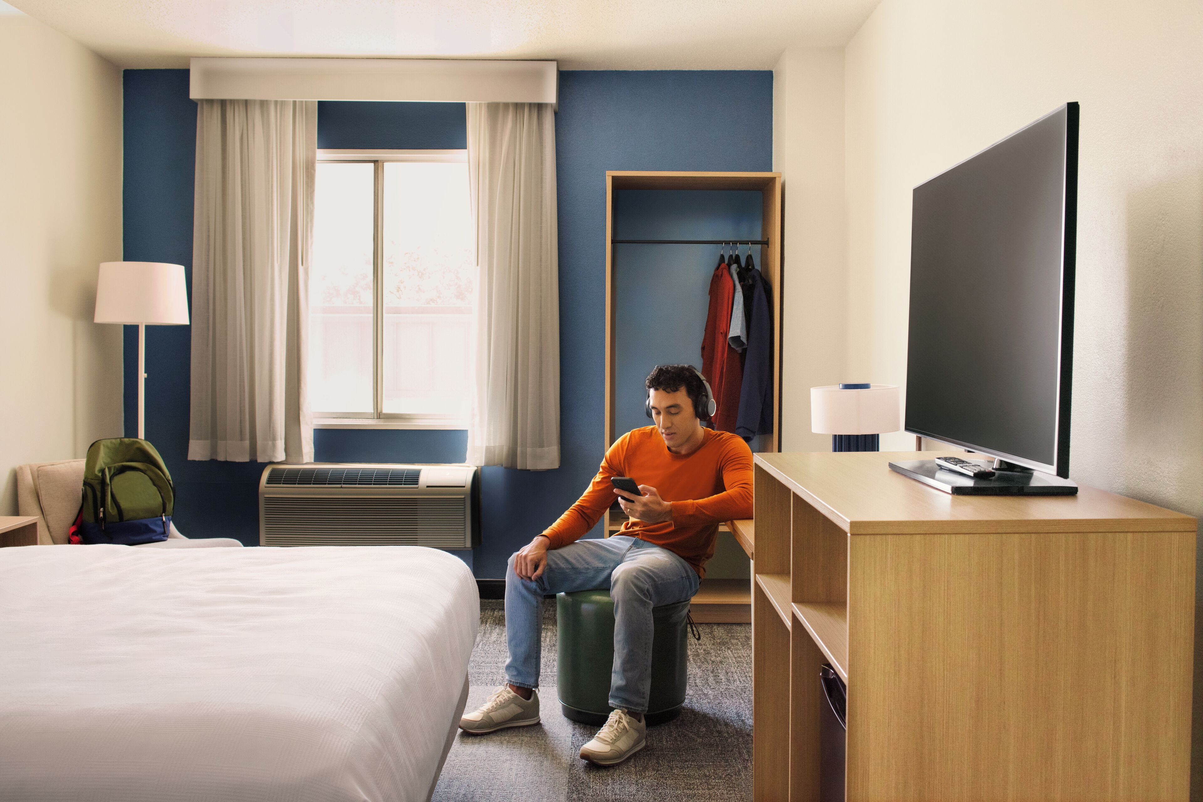 A hotel room with a bed, a desk with a TV, a chair, a lamp, a window with curtains, and a coat rack with clothes.