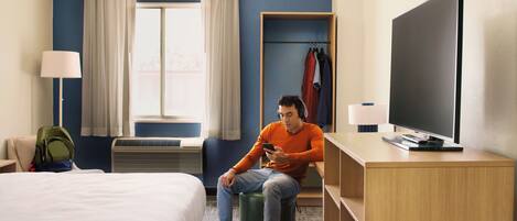 A hotel room with a bed, a desk with a TV, a chair, a lamp, a window with curtains, and a coat rack with clothes.