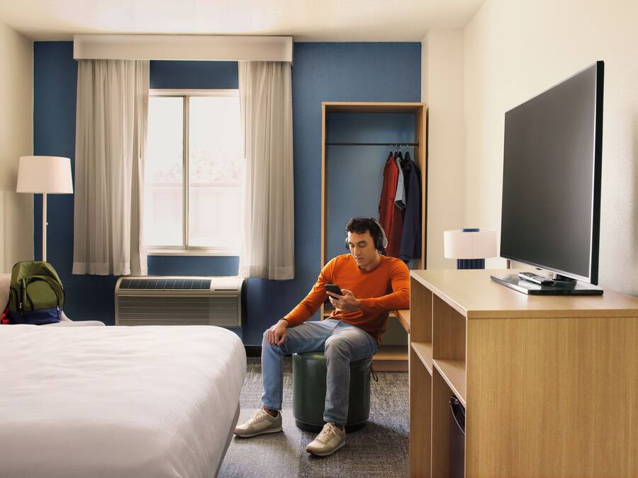 Habitación de hotel con cama, escritorio con televisor, una silla, lámpara, ventana con cortinas y perchero con ropa.