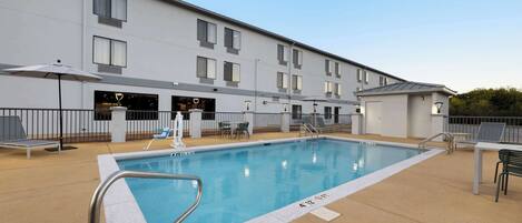 Et rektangulært pool med gelænder i metal, omgivet af en betonflisebelagt terrasse. Liggestole og parasoller, og en bygning med flere vinduer og et fladt tag i baggrunden.