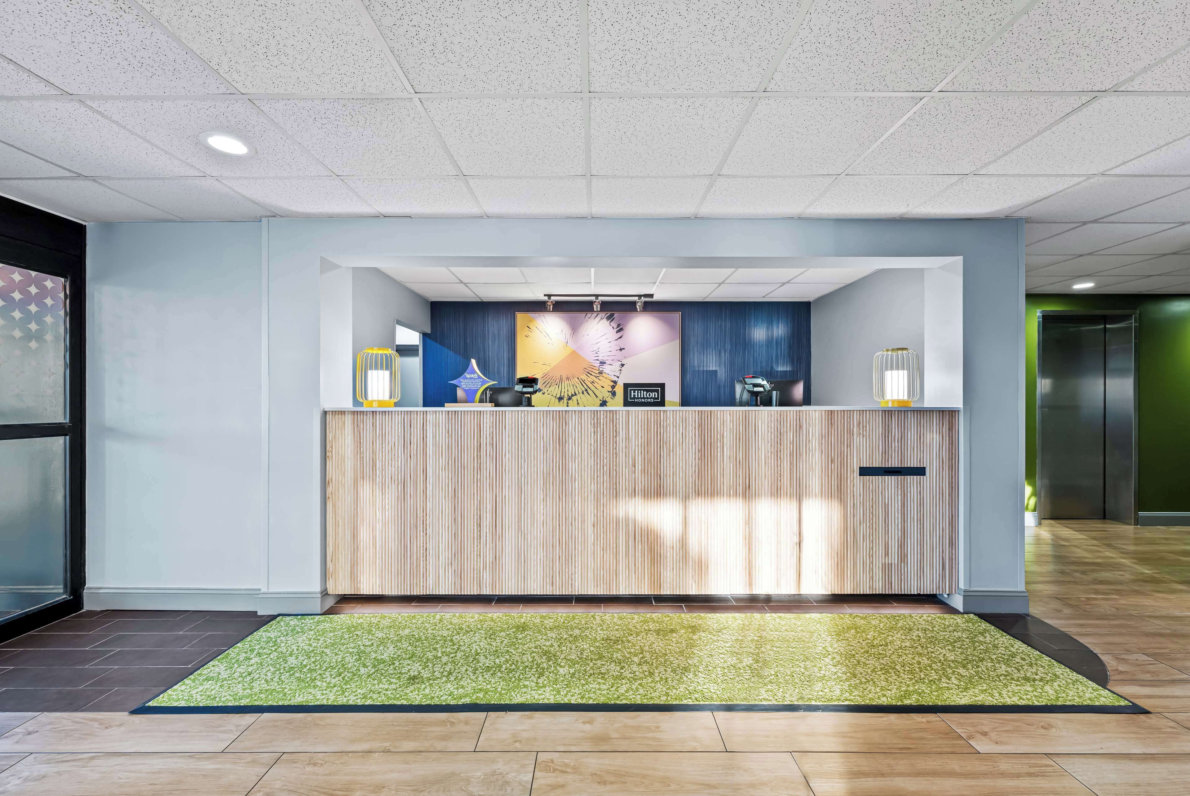 Een hotel receptie met een houten receptiebalie, een groot scherm waarop het Hilton logo staat, en een groen tapijt.