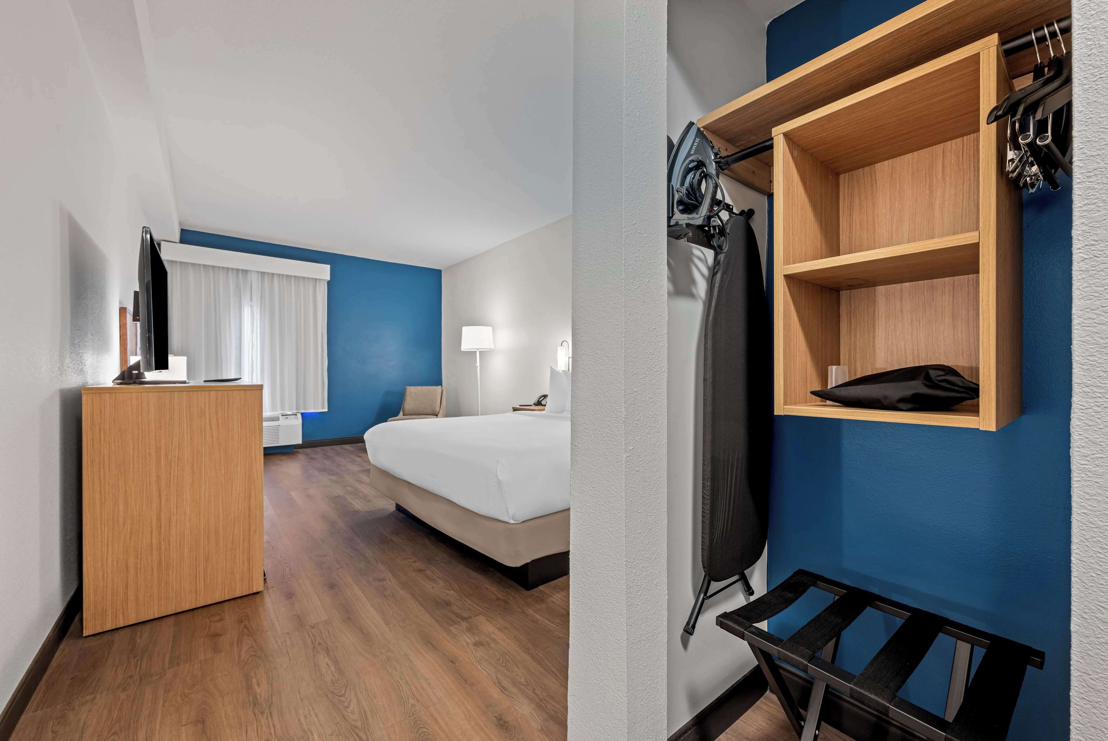 A hotel room with a wooden desk, a bed, a wardrobe, and a shelf with hanging items.