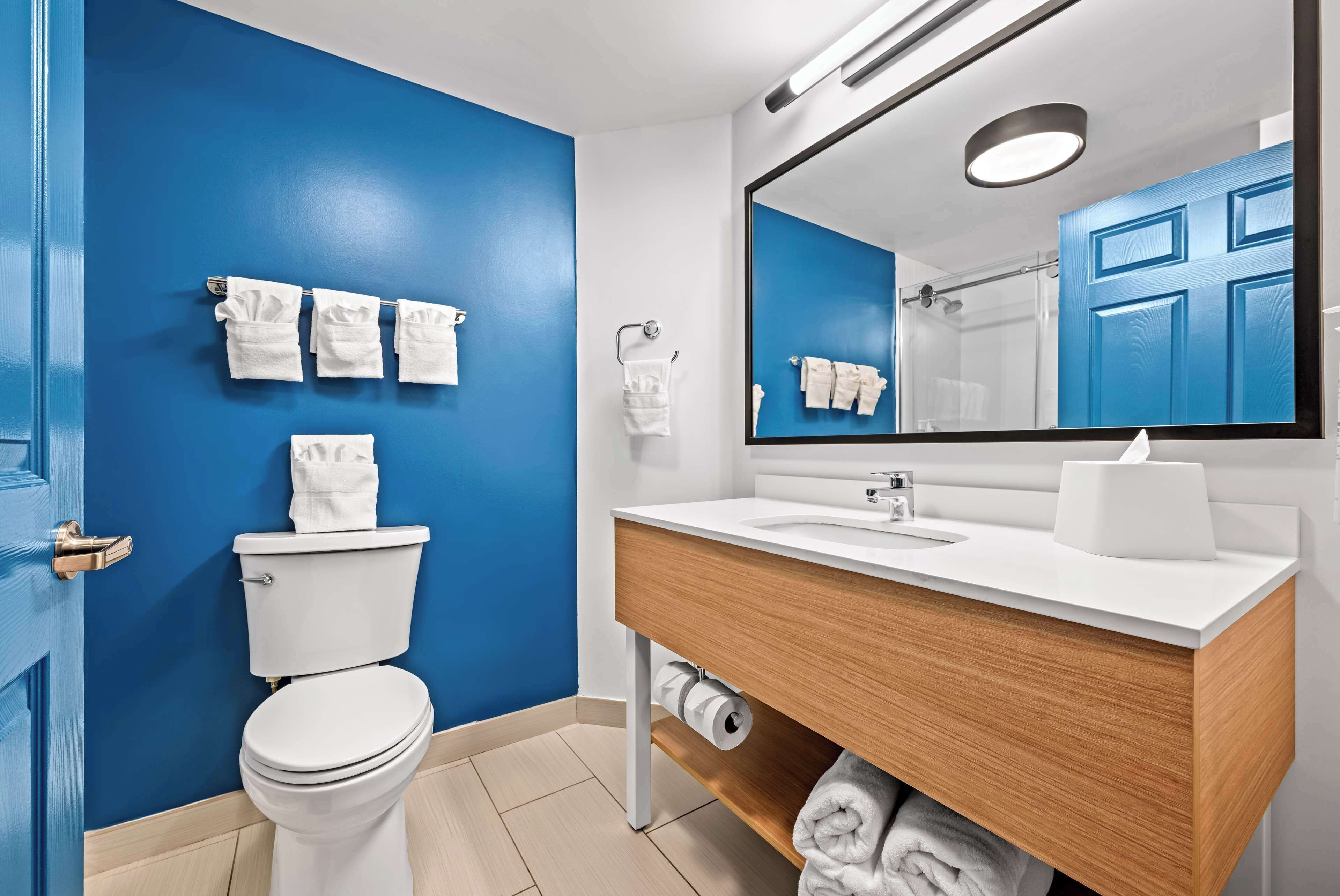 A modern bathroom with a blue accent wall, a white toilet, a wooden vanity with a mirror, and neatly arranged towels.